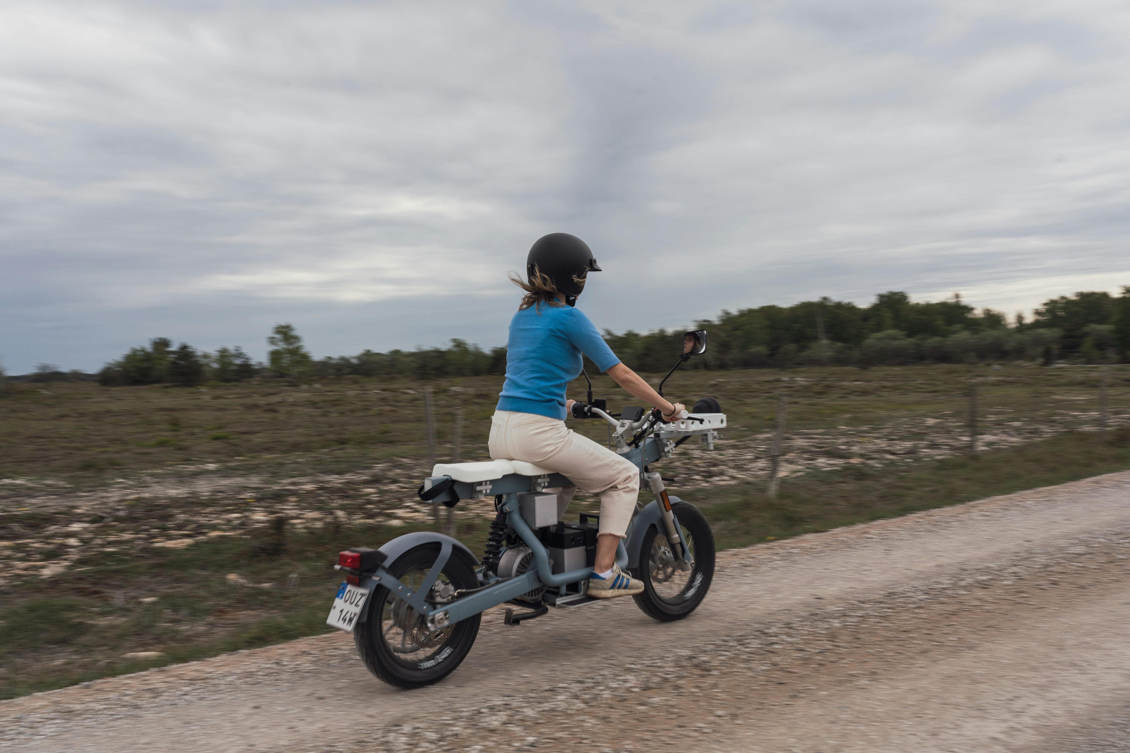 Person i blå tröja kör motorcykel på grusväg med naturlandskap i bakgrunden.