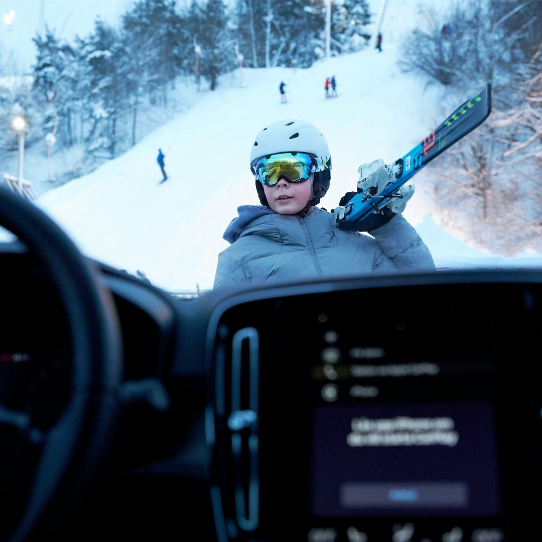 En pojke i skidhjälm och skidglasögon håller i sina skidor. Ett vinterlandskap och skidbacke i bakgrunden sett från bilens interiör.