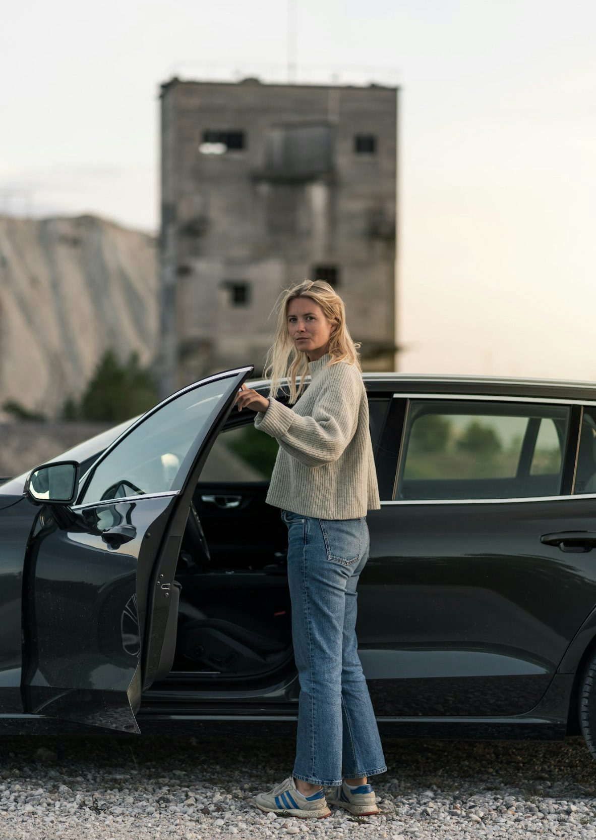 En kvinna står vid en svart Volvo med öppen dörr, i bakgrunden finns en ruin i oskärpa.