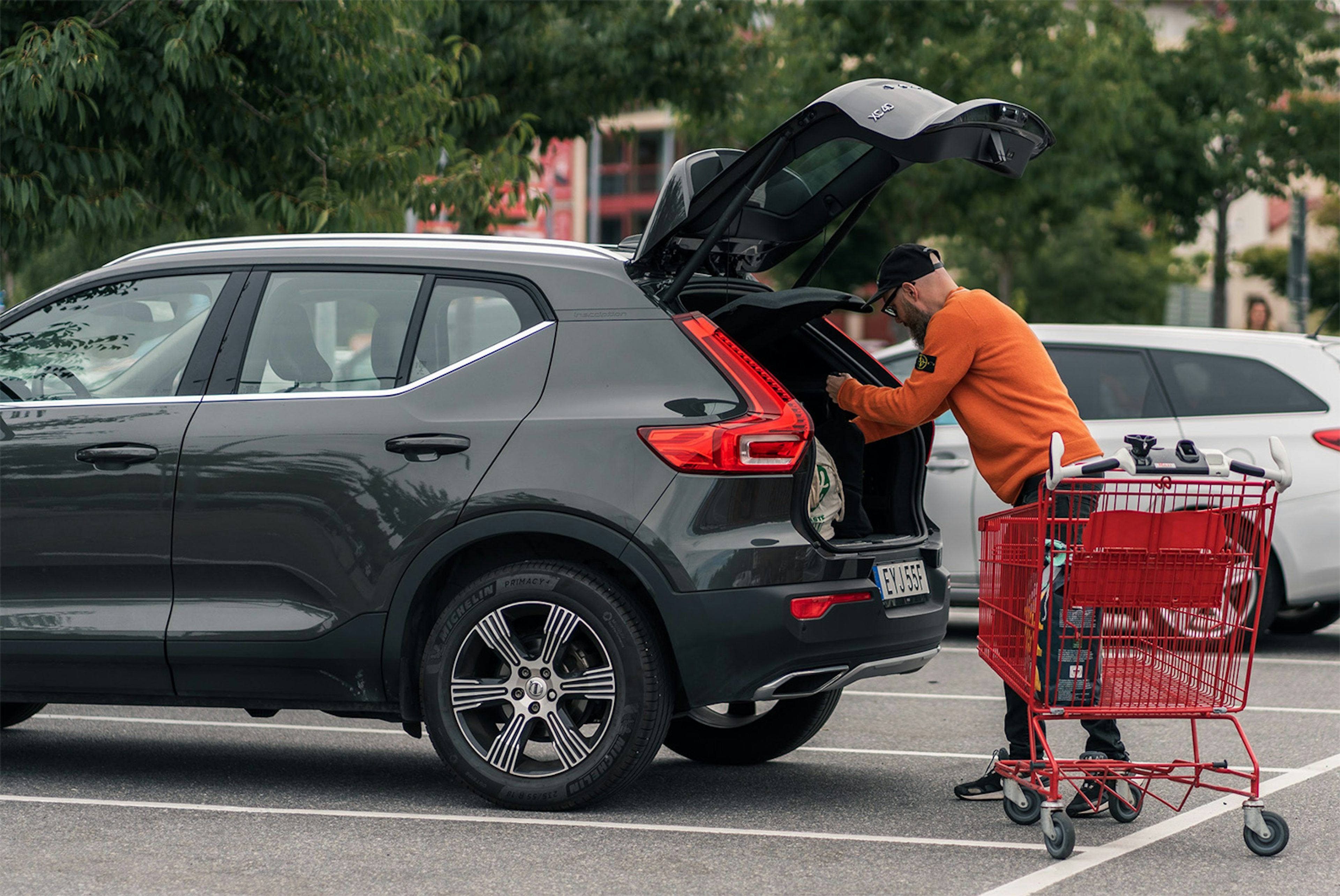 Man packar varor i bagageutrymmet på en grå SUV på parkeringsplats med shoppingvagn bredvid.