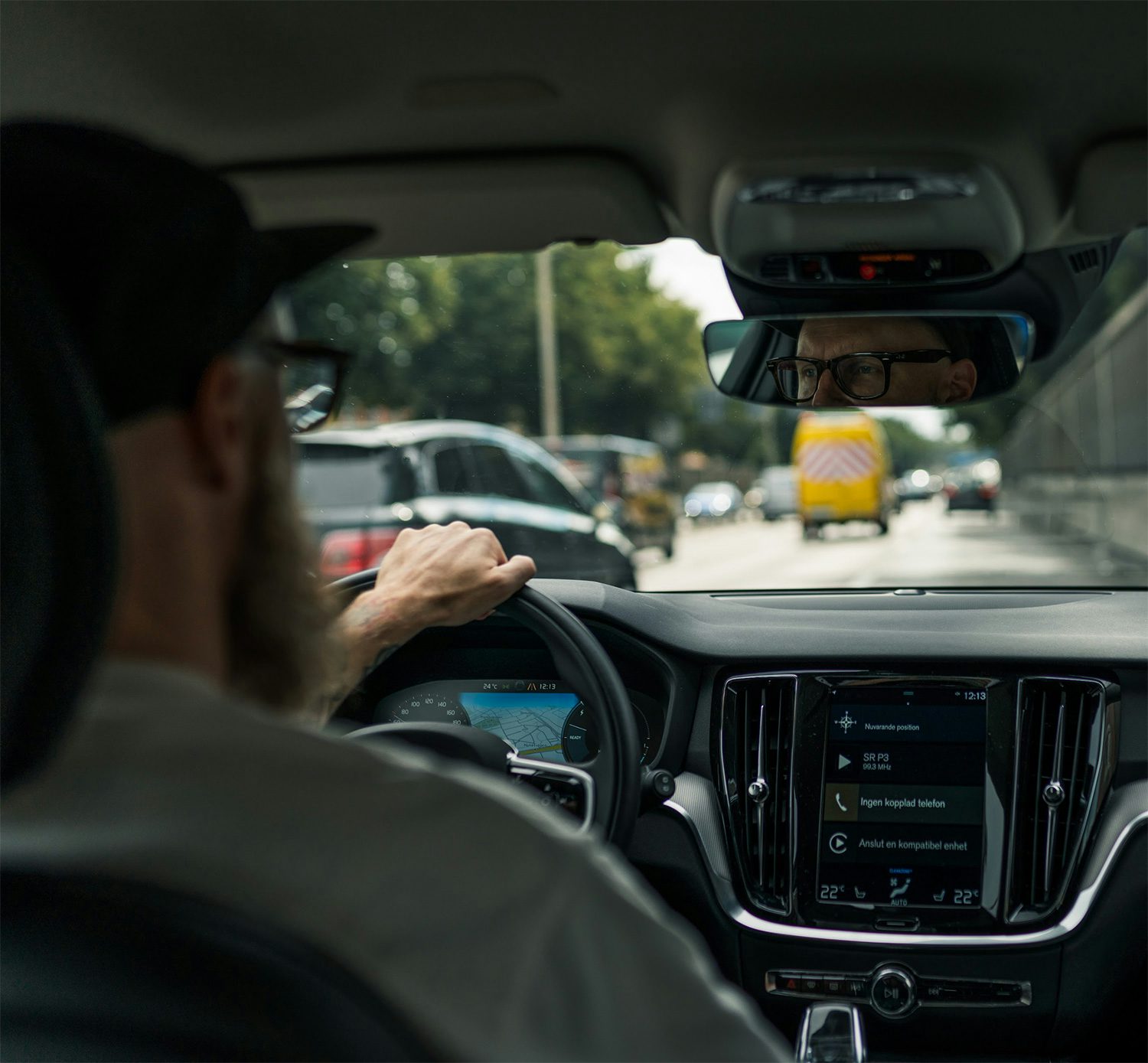 Man med skägg kör en Volvo i stadstrafik, med fokus på interiör och navigeringssystem på skärmen.