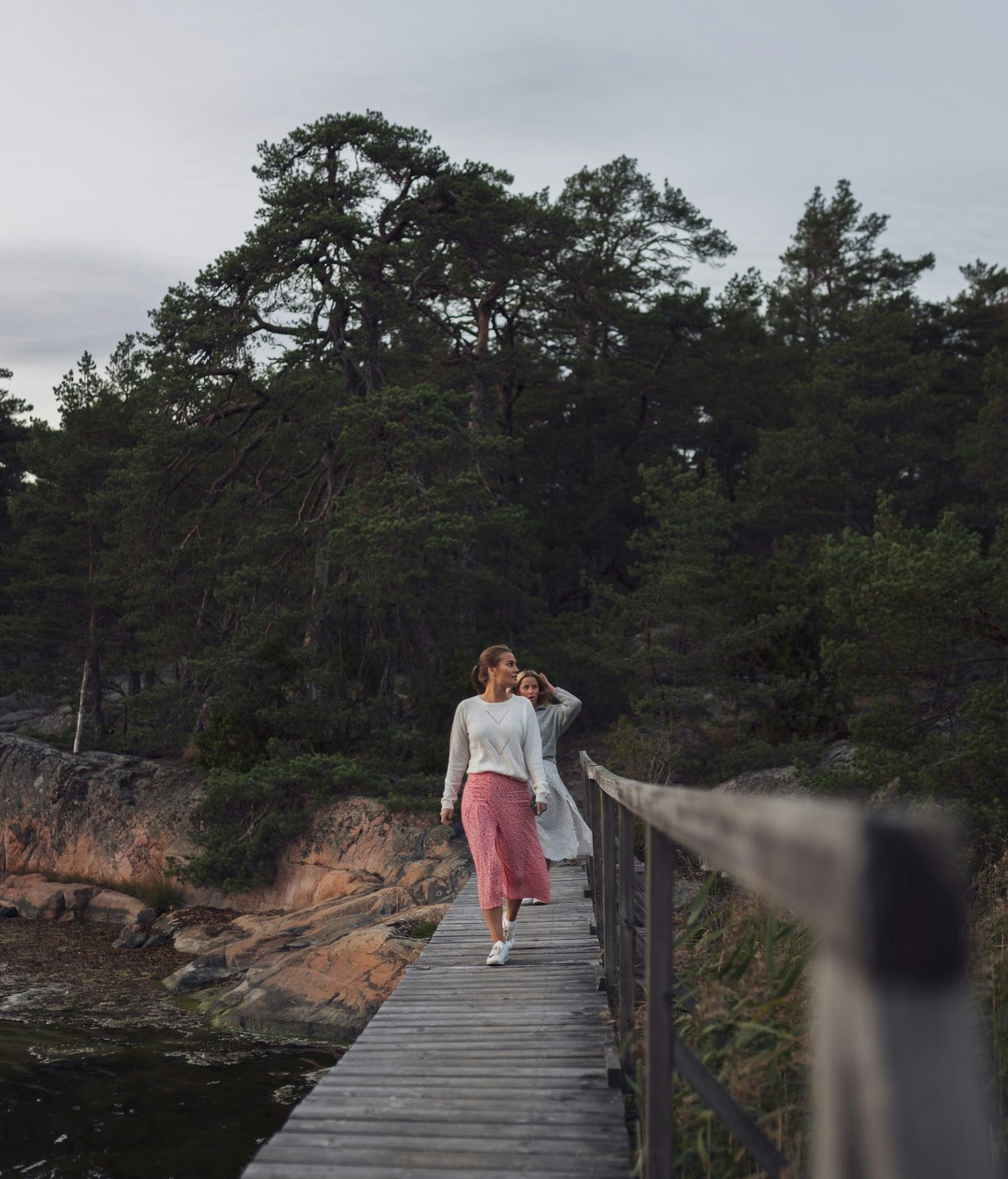 Två kvinnor går på en träbro vid havet.