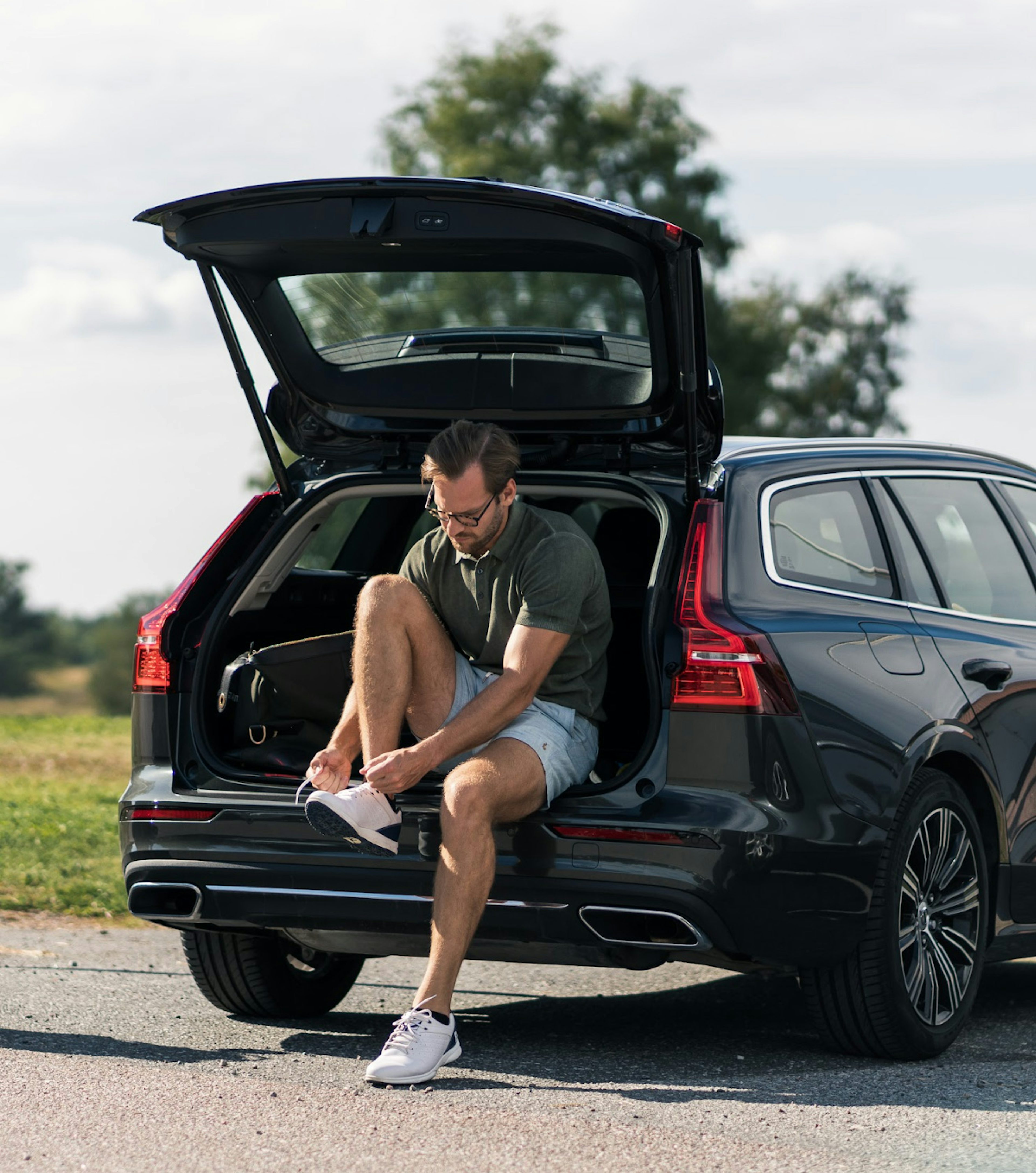 En man sitter på bakluckan av en Volvo som står parkerad i ett naturområde, han knyter sina skor.