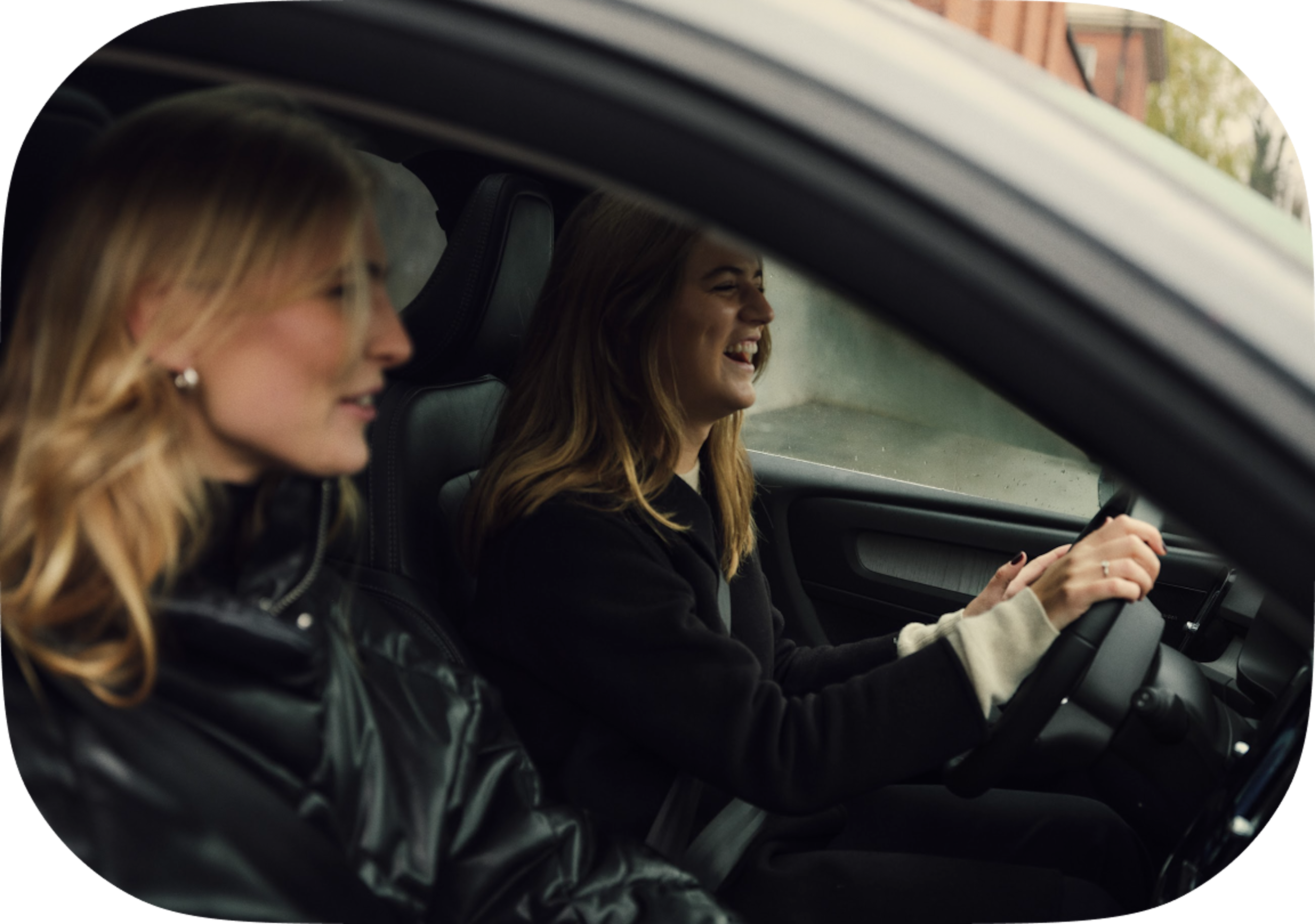 2 women sit in the front seat of a Volvo car and smile and laugh.