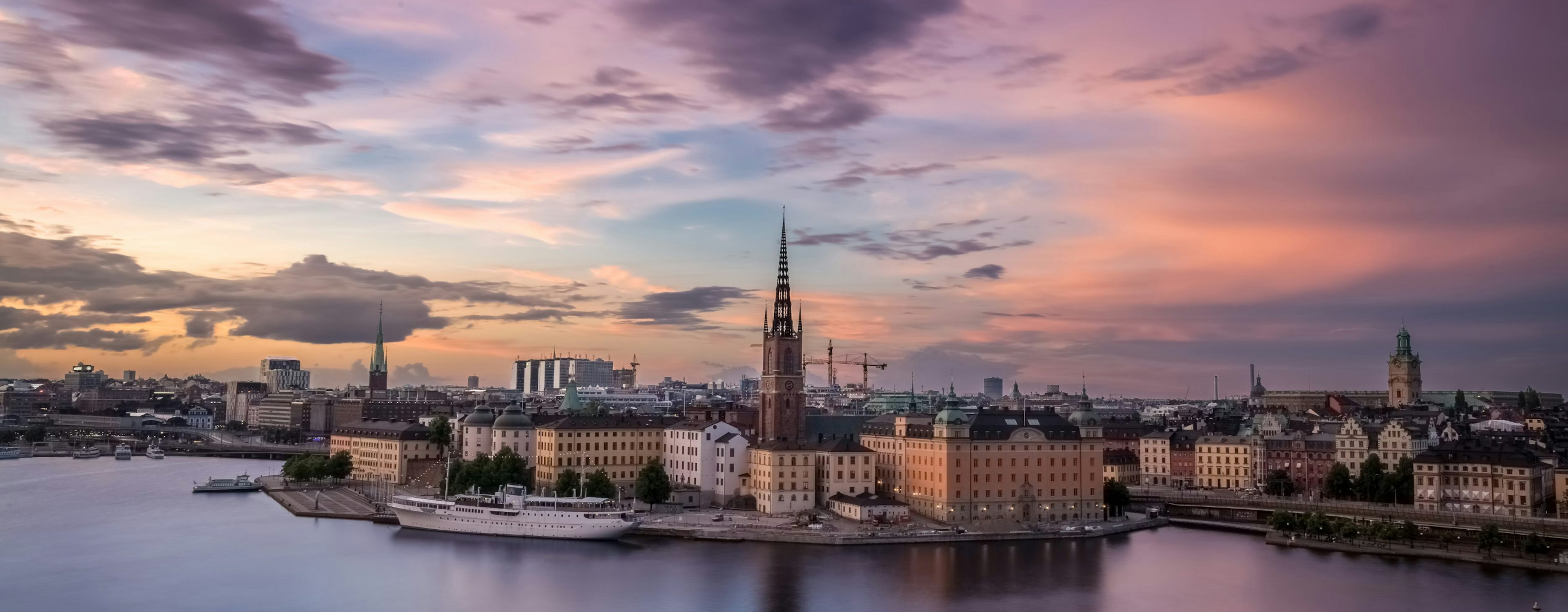 A panoramic image of the Stockholm city skyline at sunset with a pastel-colored sky and buildings reflected in the calm water