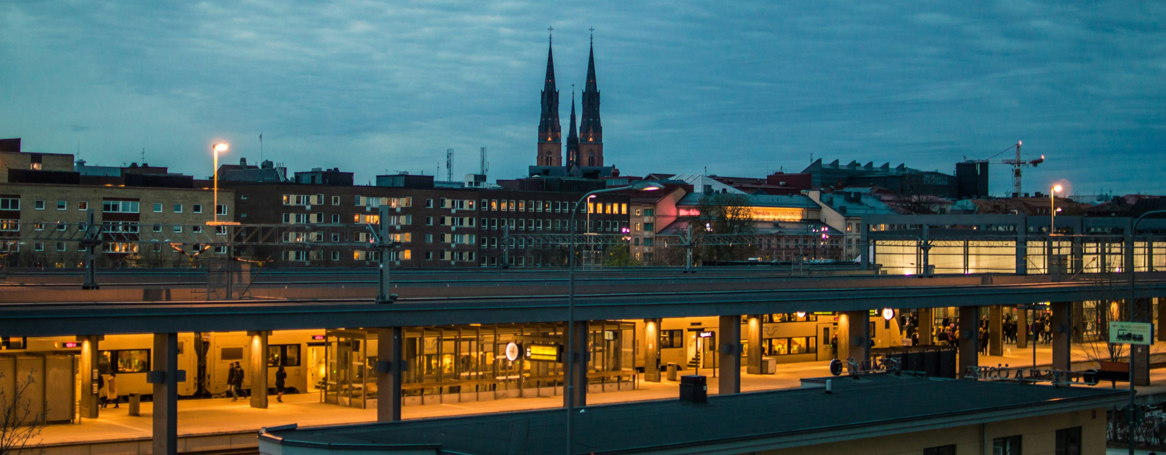 Kvällsvy över Uppsala med upplyst tågstation i förgrunden och den ikoniska Uppsala domkyrka i bakgrunden